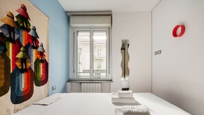 A hotel room with a bed, a large abstract painting, a window with a view, and a red circular object on the wall.