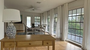 Tilava ruokailutila, jossa on puinen pöytä, tuolit ja kattotuuletin. Suuret ikkunat, joissa on valkoiset verhot, päästävät runsaasti luonnonvaloa sisään ja tarjoavat näkymät ulos.
