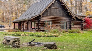 Une cabane en rondins surmontée d’un toit à forte pente, entourée d’un jardin bien entretenu et bordée par une forêt.