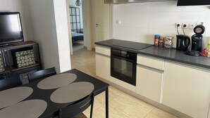 Une cuisine moderne avec un plan de travail noir, des armoires blanches et un four encastré. On y trouve un coin repas avec une table et des chaises, ainsi qu’un téléviseur posé sur un meuble.