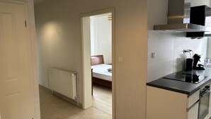 Une cuisine compacte équipée d’une cuisinière, d’un four et d’un petit plan de travail. On aperçoit un radiateur sur le sol, et un lit est visible à travers une porte ouverte donnant sur la pièce adjacente.