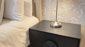 A neatly made bed with white linens and pillows, a black nightstand with a lamp, and a patterned wallpaper background.