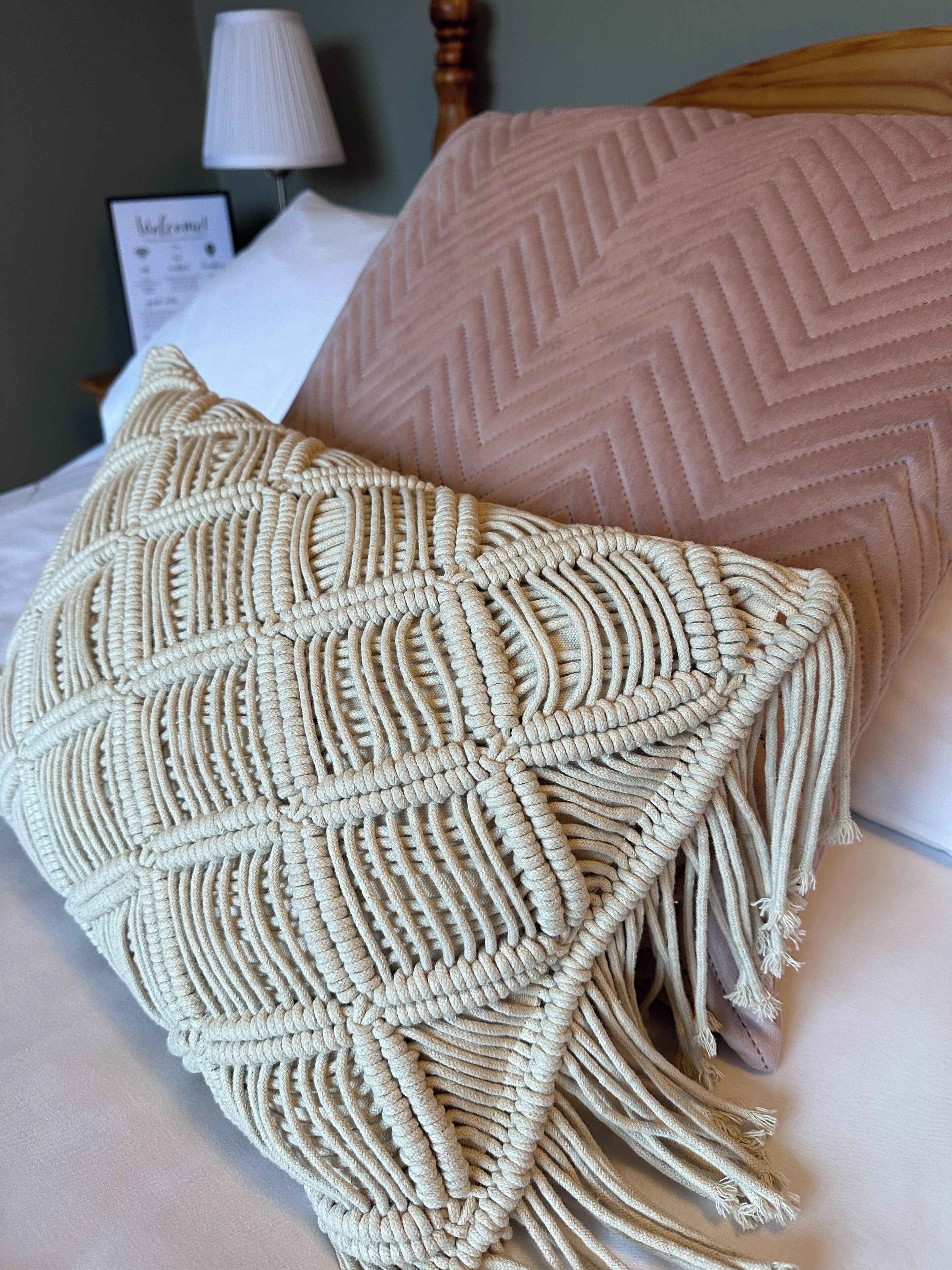 A bed with a macrame throw blanket and a pink quilted pillow.