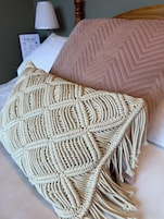 Una cama con un plaid de macramé y una almohada acolchada rosa.