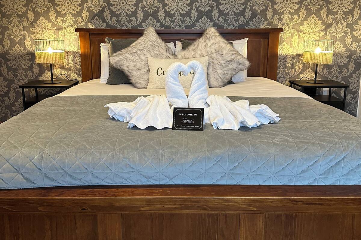 A neatly made bed with a quilted grey bedspread, two fluffy pillows, a heart-shaped towel arrangement, and a welcome sign on the bed.