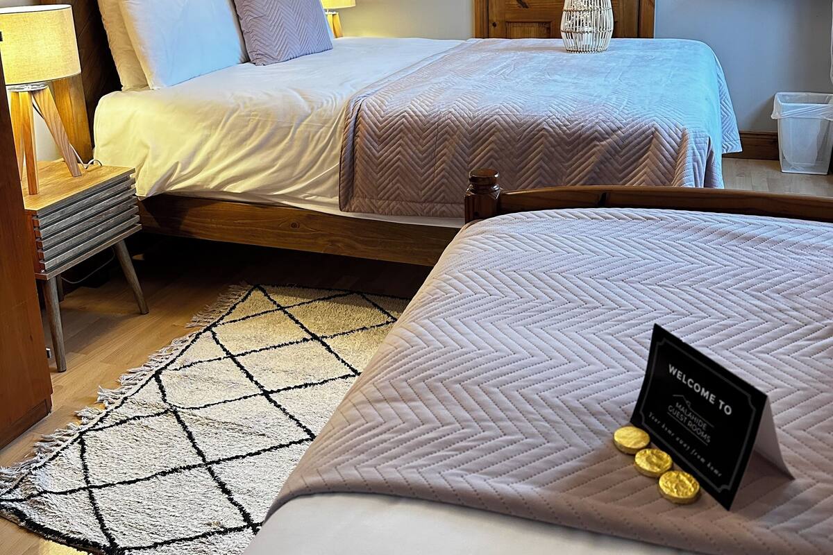 A two-bed room with a wooden headboard, a bedside table with a lamp, a wall-mounted shelf with towels, a wooden wardrobe, and a welcome sign on the bed.