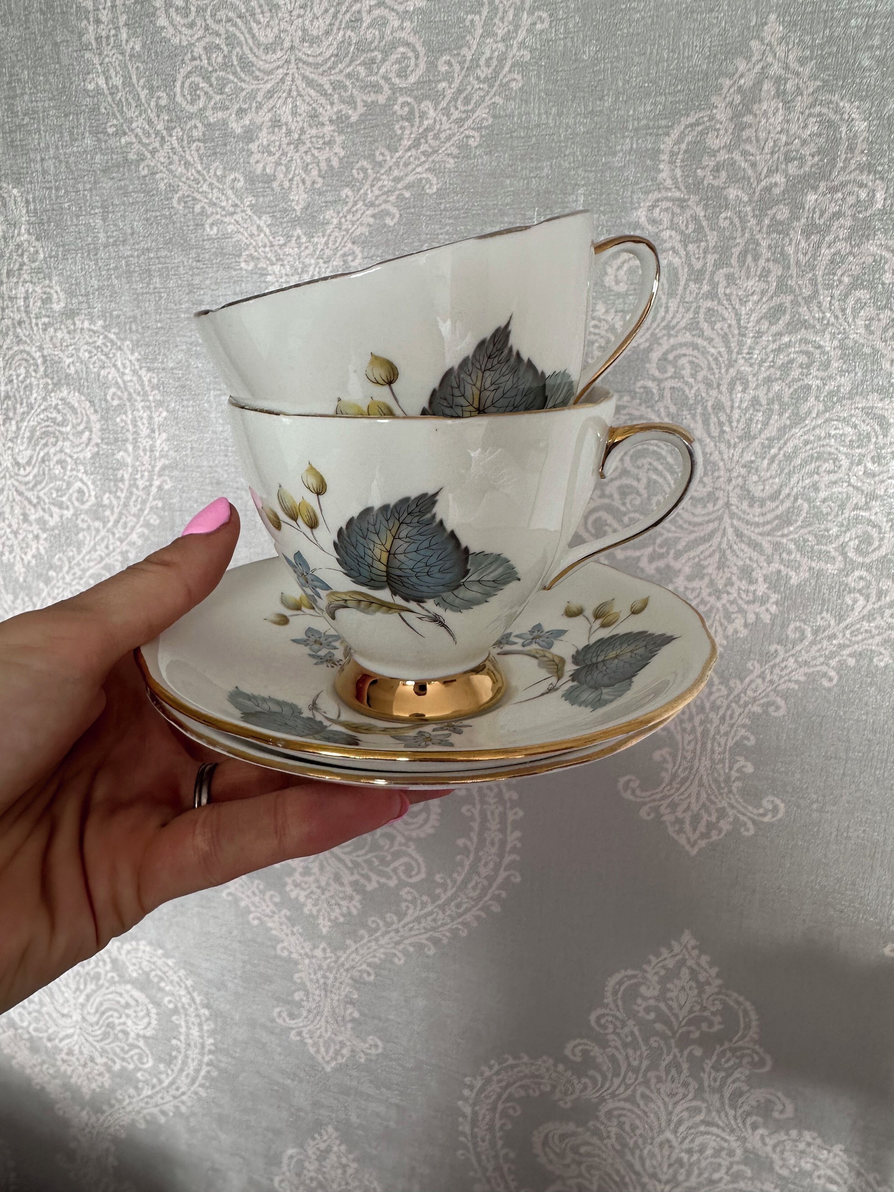 A hand holding a white teacup with floral designs and a matching saucer, with a gold rim.