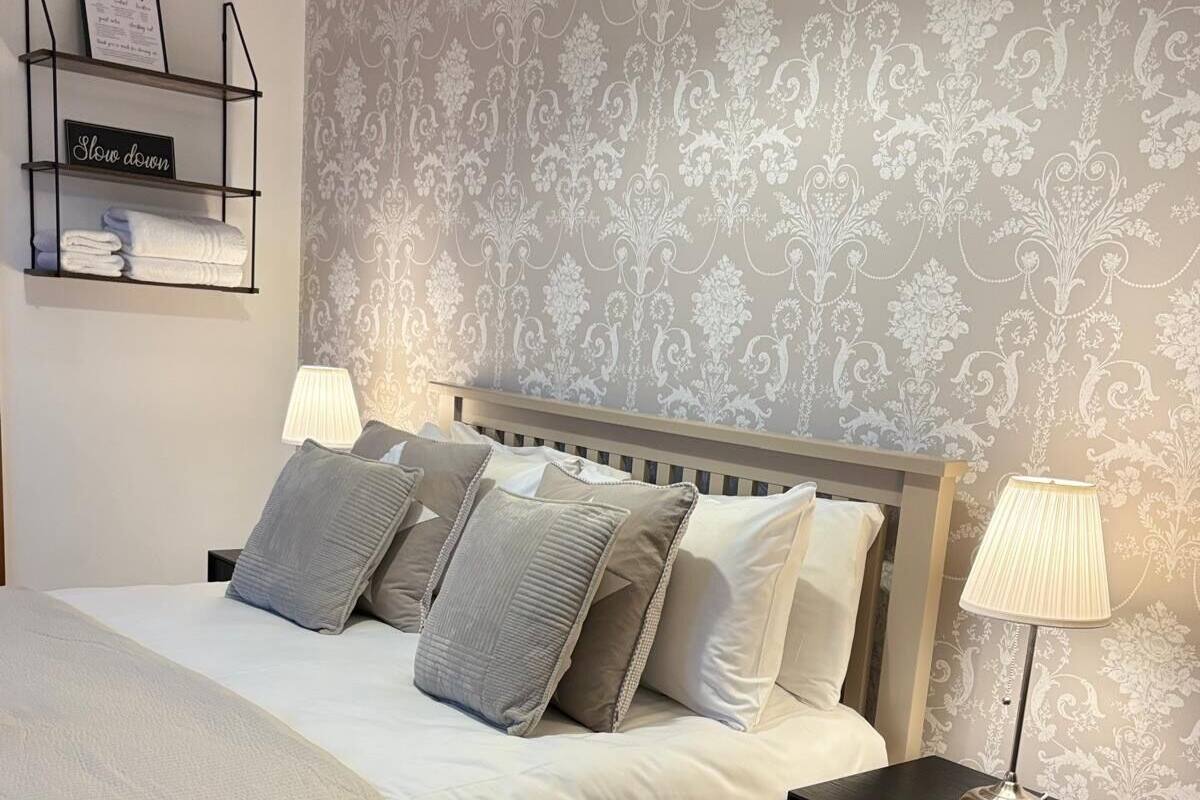 A neatly made bed with a white comforter and several pillows, a bedside table with a lamp, a patterned wallpaper, and a small shelf with a welcome sign and a 'Slow down' sign.
