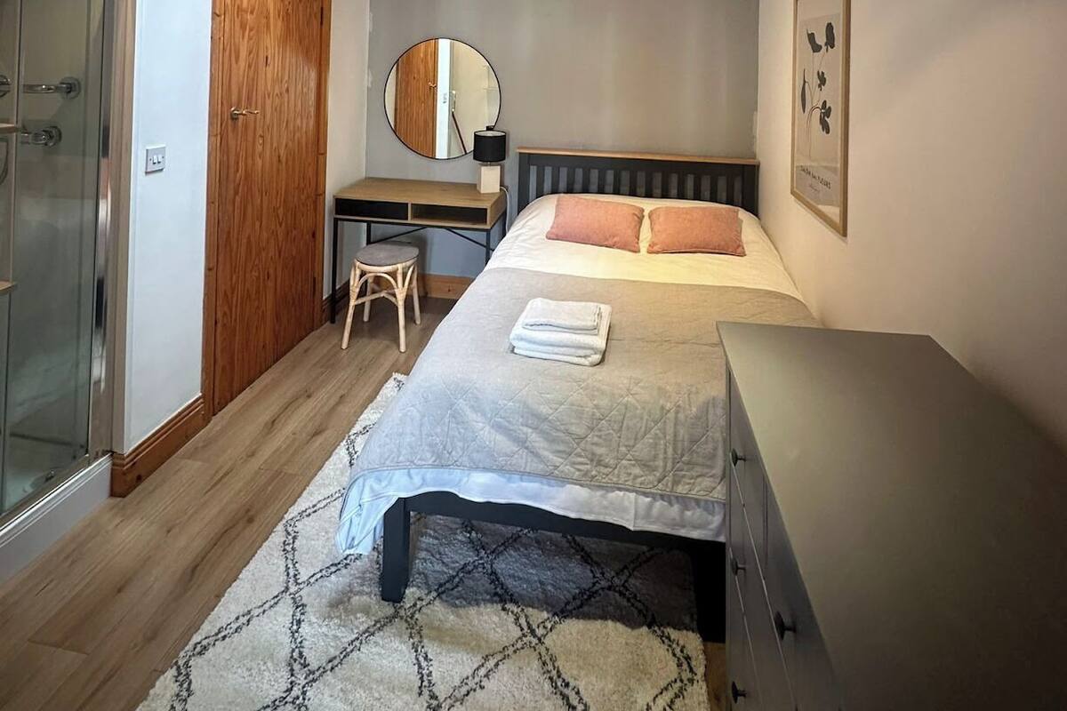 A bedroom with a bed, a desk, a chair, a mirror, and a shower.