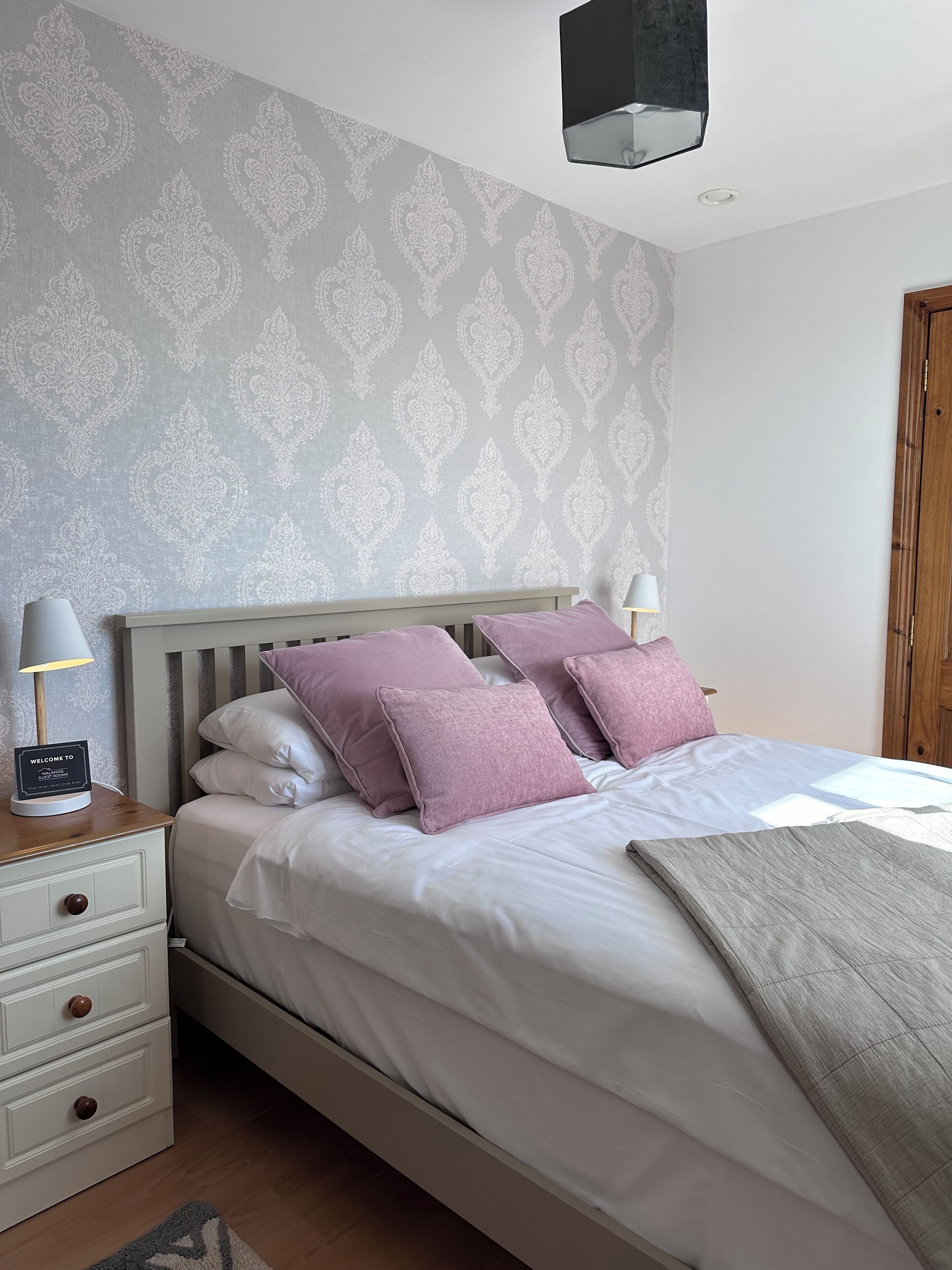 A neatly made bed with white and purple bedding, a bedside table with a lamp, and a welcome sign.