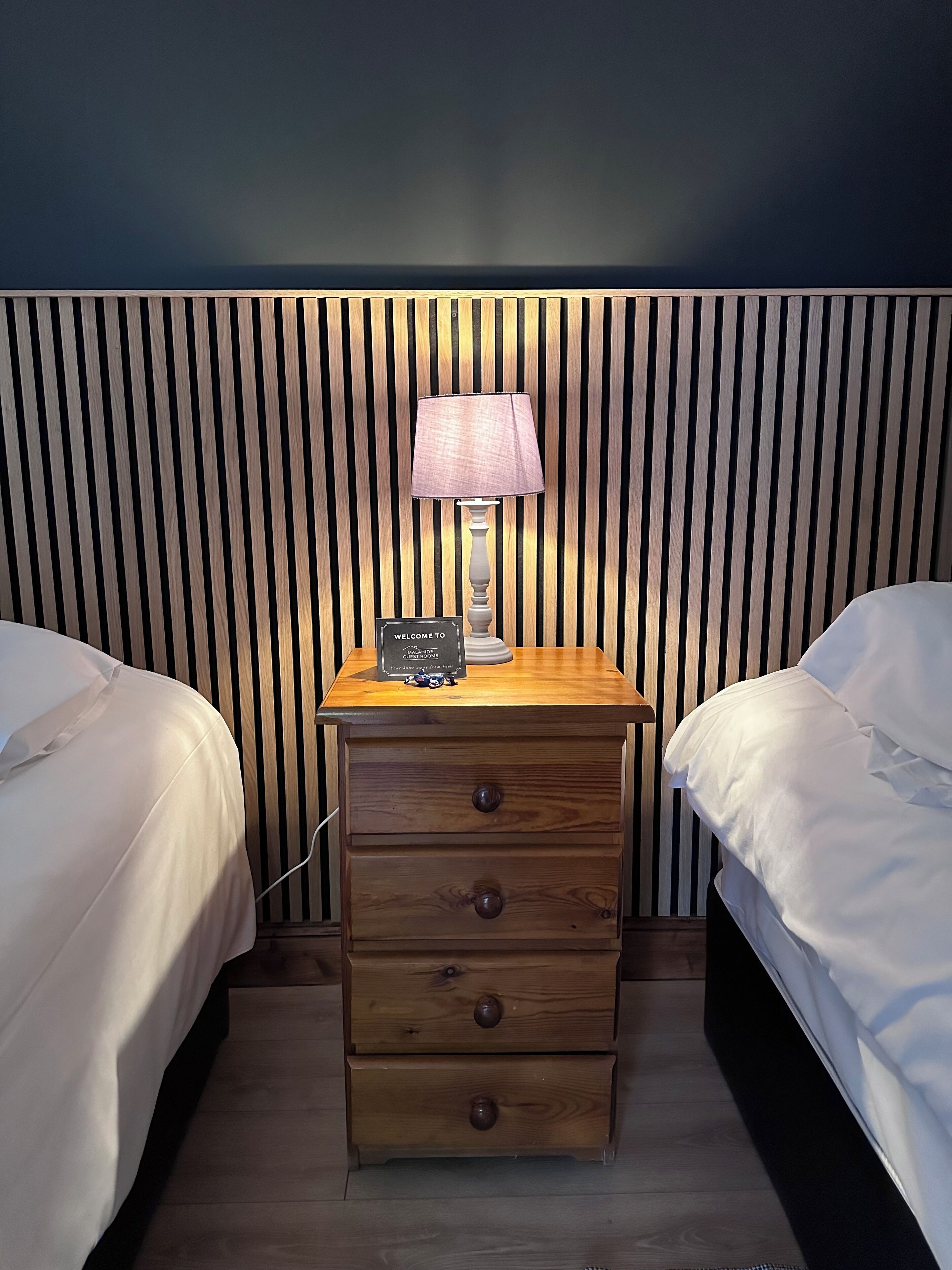 Hotellihuone, jossa on kaksi erillistä sänkyä, puinen yöpöytä lampun kanssa ja kyltti, josta ilmenee, että kyseessä on vierashuone.