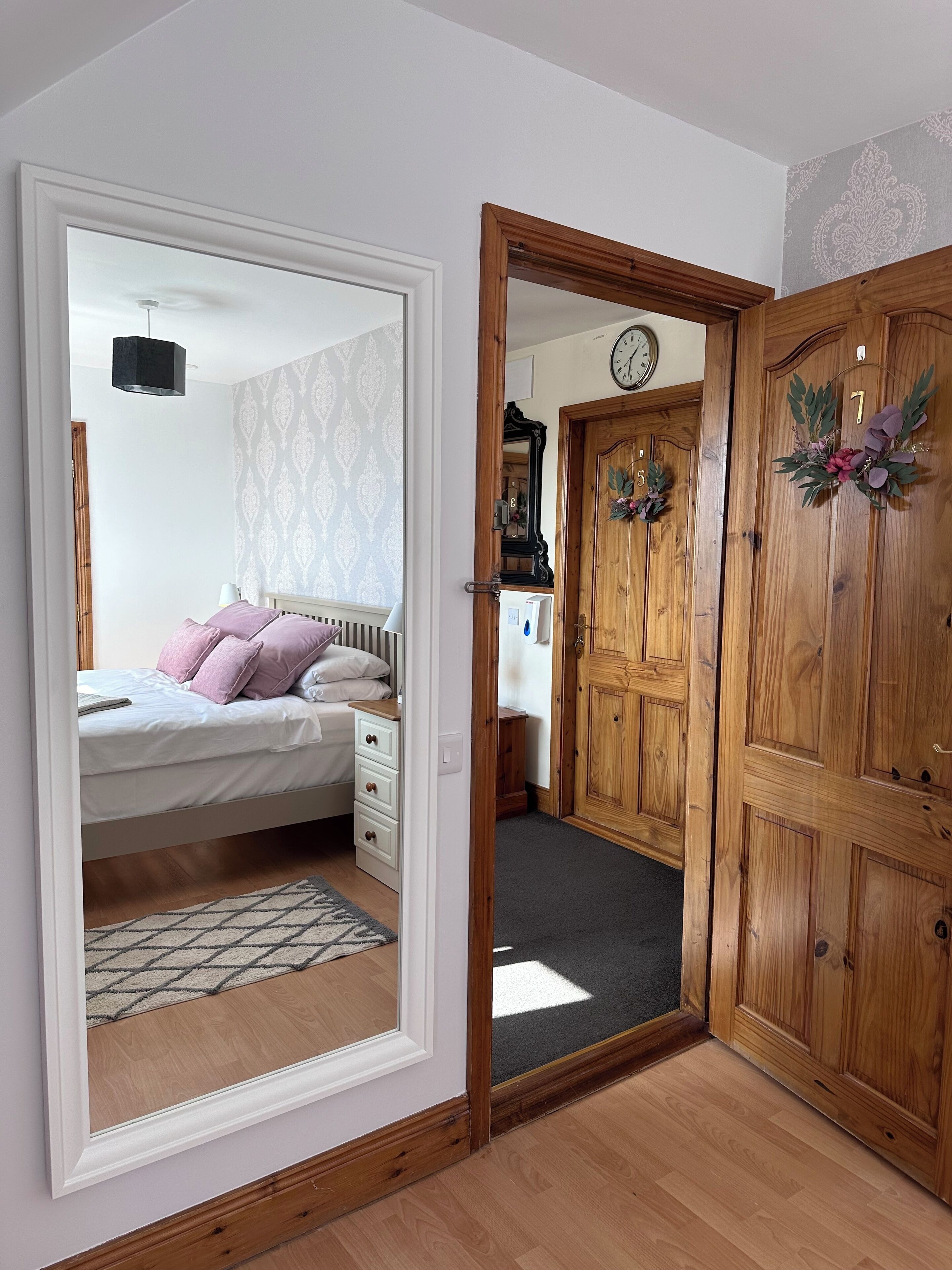 Slaapkamer met een bed, een kaptafel en een spiegel die een hal met houten deuren en een klok weerkaatst.