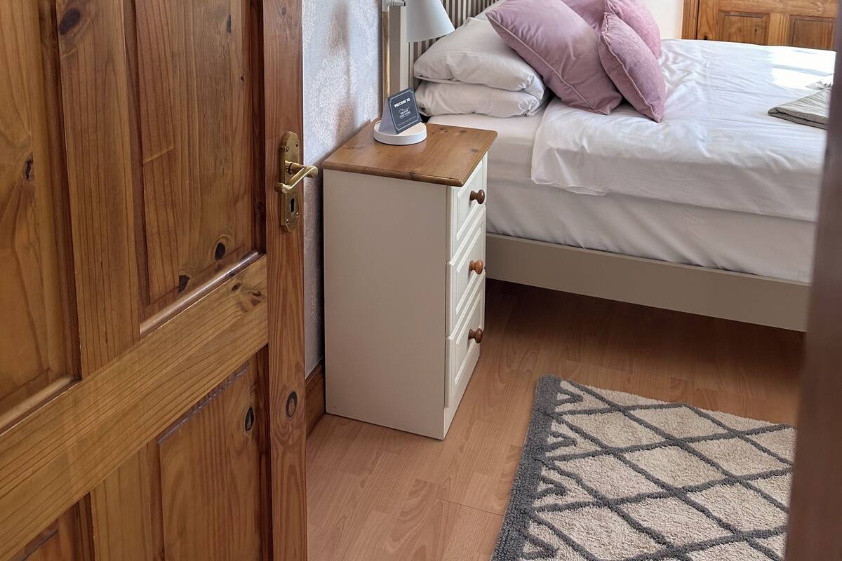 A bedroom with a wooden wardrobe, a bedside table with a lamp, a bed with white and pink bedding, and a geometric rug.