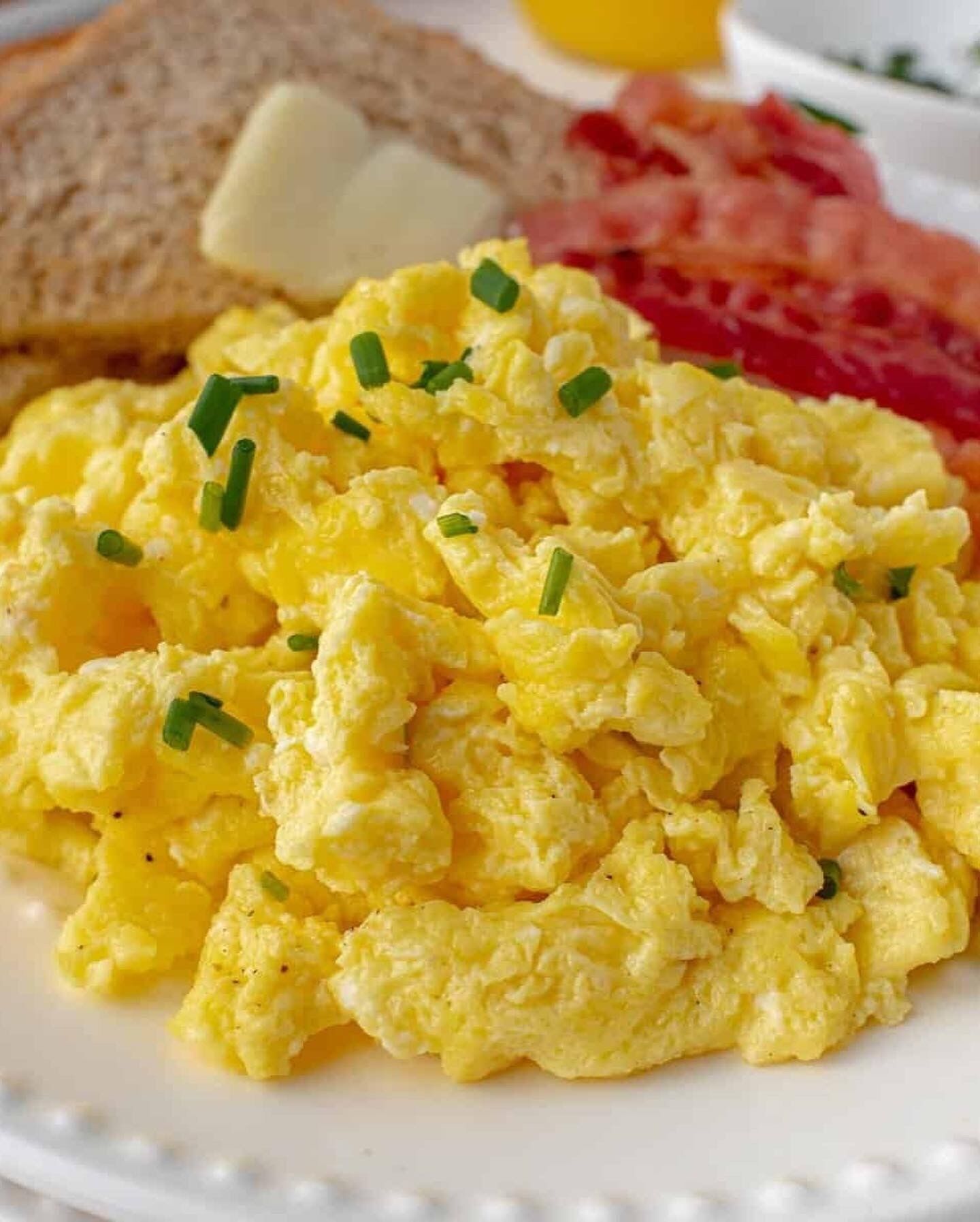Scrambled eggs garnished with chives, bacon, toast, and a glass of orange juice on a plate.
