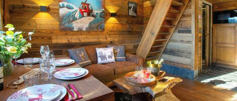 Ein gemütliches Blockhaus-Interieur mit einem Holzespeisetisch, gedeckten Tisch, einer Couch mit Zierkissen, einem Bild vom Sessellift und einer Treppe.
