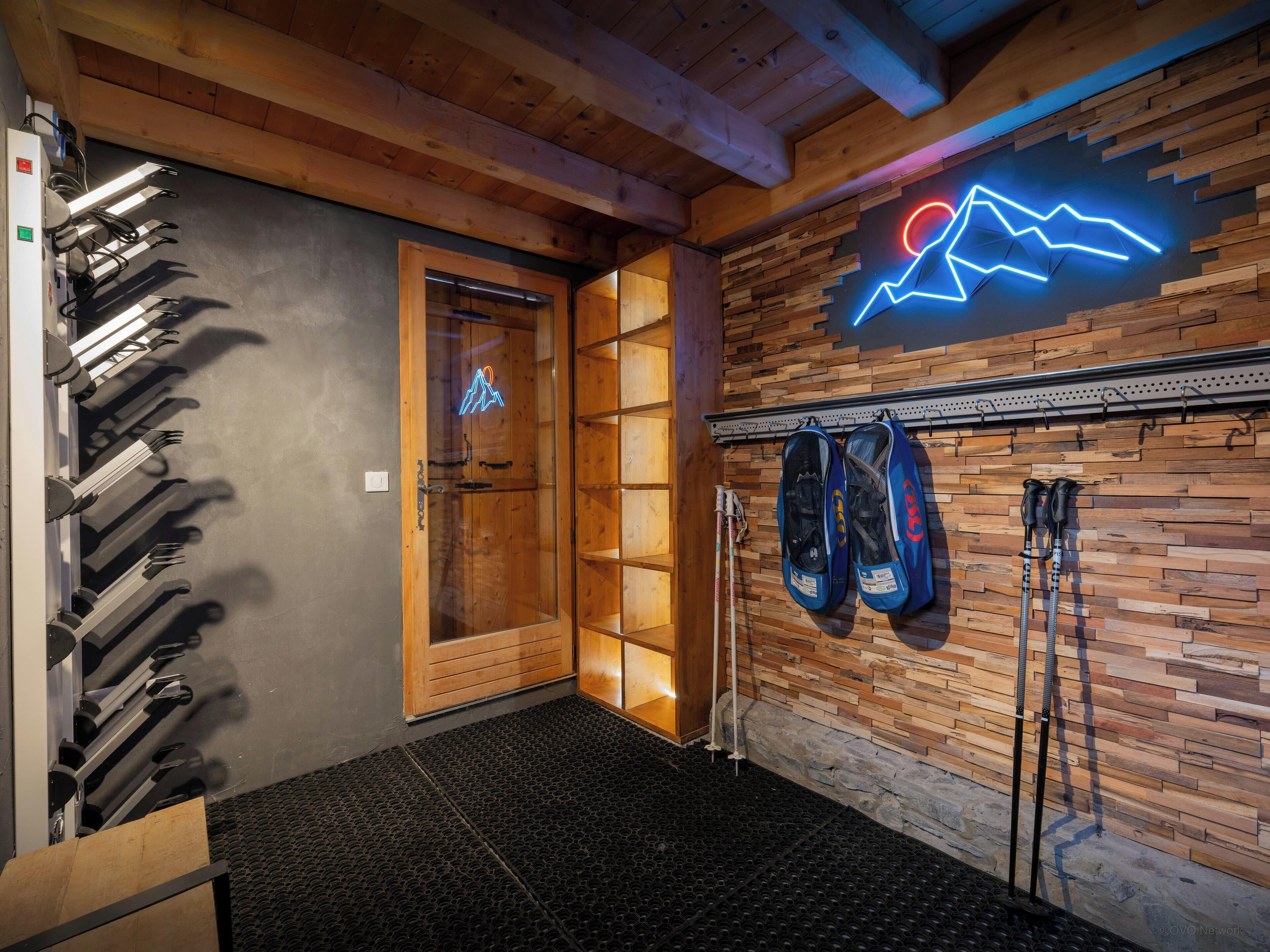 A ski lodge interior with a wooden wall, a neon mountain design, and ski equipment on racks.