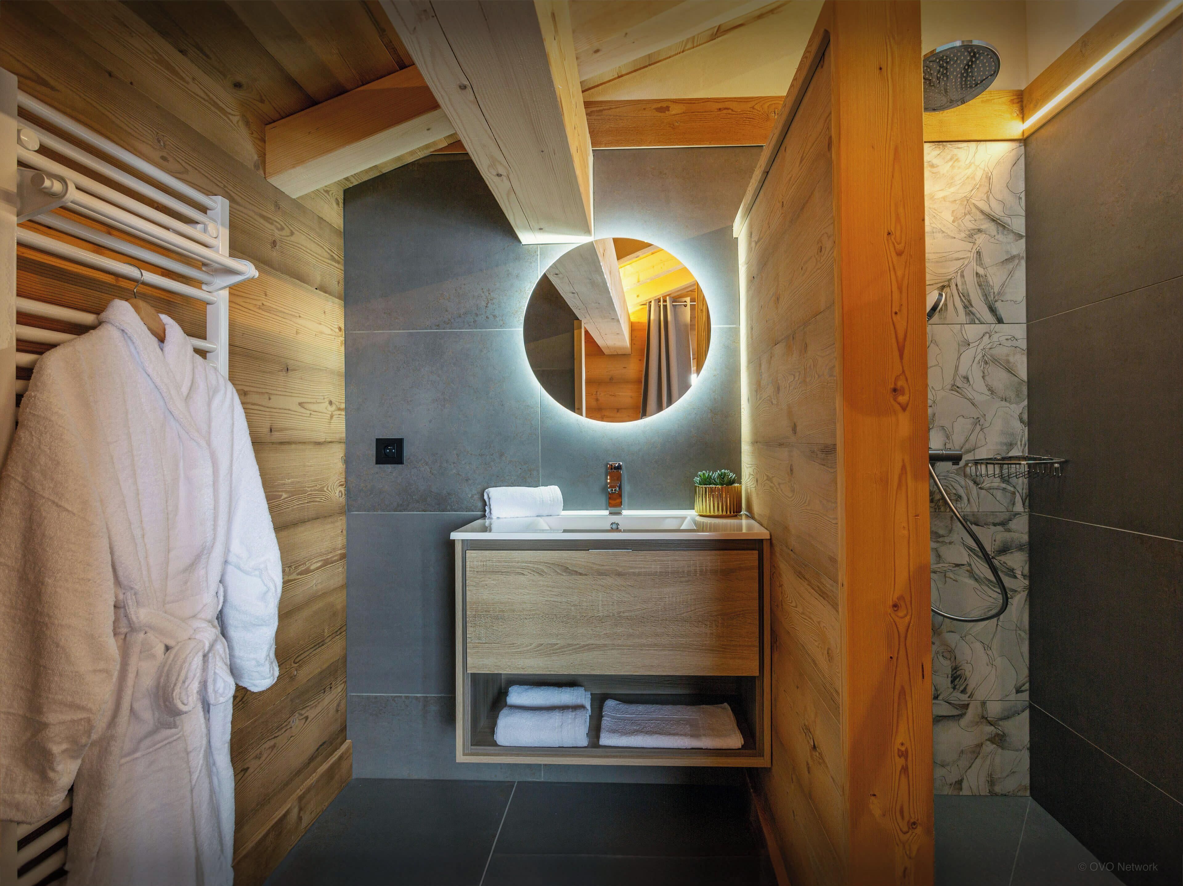 A modern bathroom with a wooden vanity, a round mirror, and a shower area.