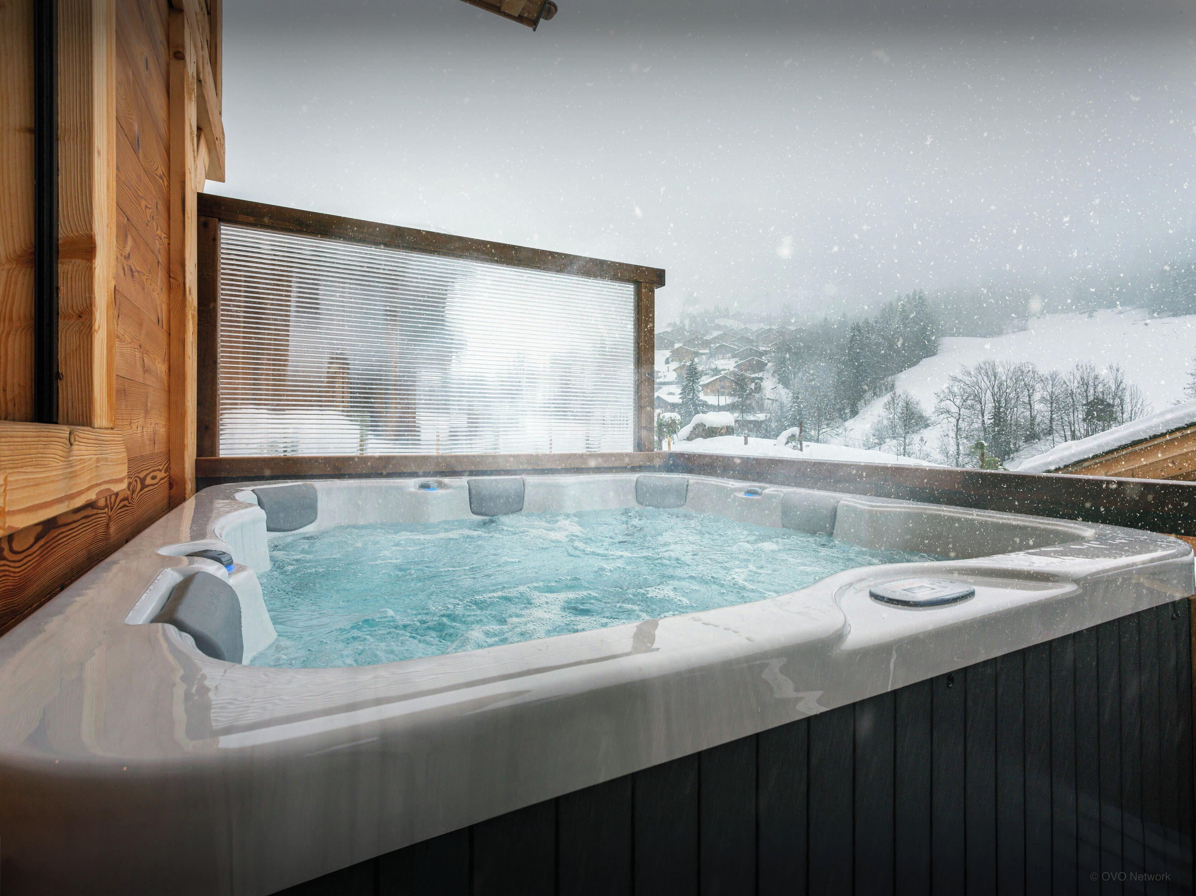 A hot tub with a wooden exterior, a window with blinds, and a snowy landscape view.