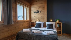 Ein gemütliches Schlafzimmer mit einem hölzernen Kopfende, einem großen Bett mit grauer Bettdecke, zwei Kissen, einem Nachttisch mit Lampe und einem Fenster mit Vorhängen.