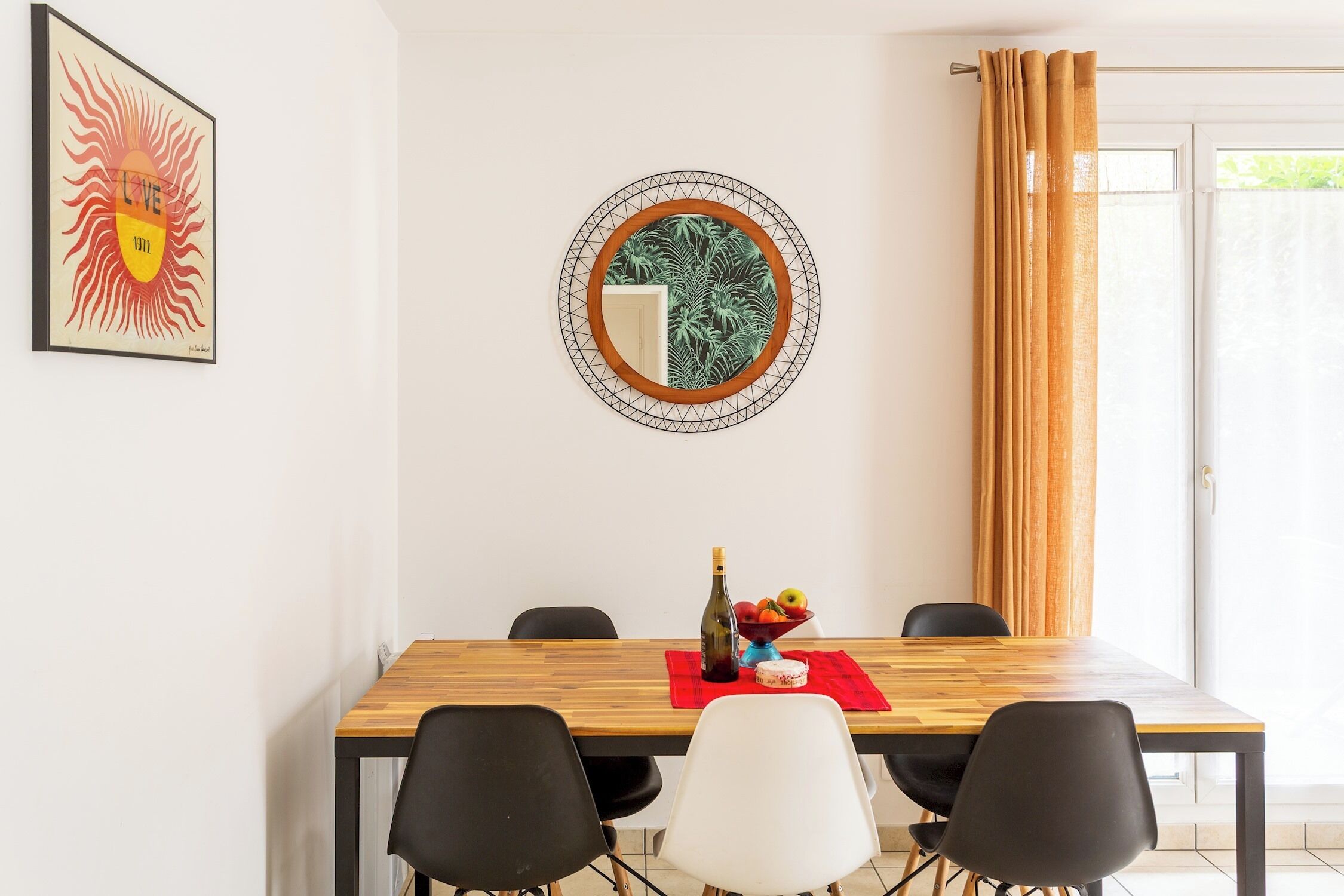 Un coin repas avec une table en bois, des chaises noires et blanches, un set de table rouge, une bouteille et un saladier rempli de fruits.