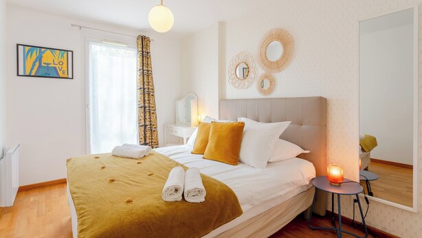 A neatly made bed with a brown blanket and two rolled towels, a bedside table with a lamp, and a mirror on the wall. - Happy Stay Disneyland Paris - Garden Apartment + Parking (Montévrain)