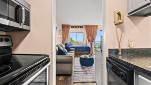 Cocina compacta con microondas, estufa y lavavajillas. Se ve un dormitorio con cama, sofá azul y cortinas a través de una puerta abierta.