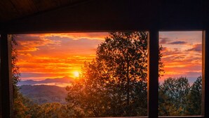 Eine Holzterrasse mit einem runden Tisch und Stühlen, mit Blick auf einen Sonnenuntergang über Bäumen und Bergen.