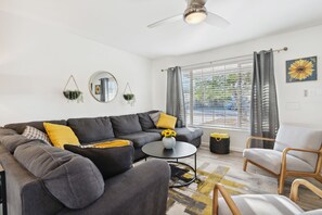 A modern living room with a grey sofa, a round coffee table, and a wooden armchair. There is a large window with curtains, a ceiling fan, and a sunflower painting on the wall.
