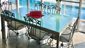 A glass-top dining table with metal chairs, a red placemat, and a view of a swimming pool and greenery through large windows. - Centrally located ground floor of 2 storey Artist's modern studio house. (Port Douglas)