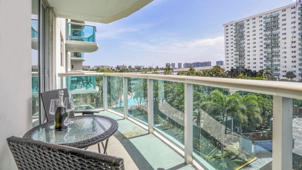 A balcony with a glass table and chairs, overlooking a swimming pool and palm trees. - Amazing Condo 3 min to the Beach (Sunny Isles Beach)