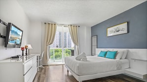 A modern bedroom with a large bed, a desk, and a view of a cityscape through the window. - Amazing Condo 3 min to the Beach (Sunny Isles Beach)