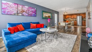 A modern living room with a blue sectional sofa, a round coffee table, and a large abstract painting on the wall. - Amazing Condo 3 min to the Beach (Sunny Isles Beach)