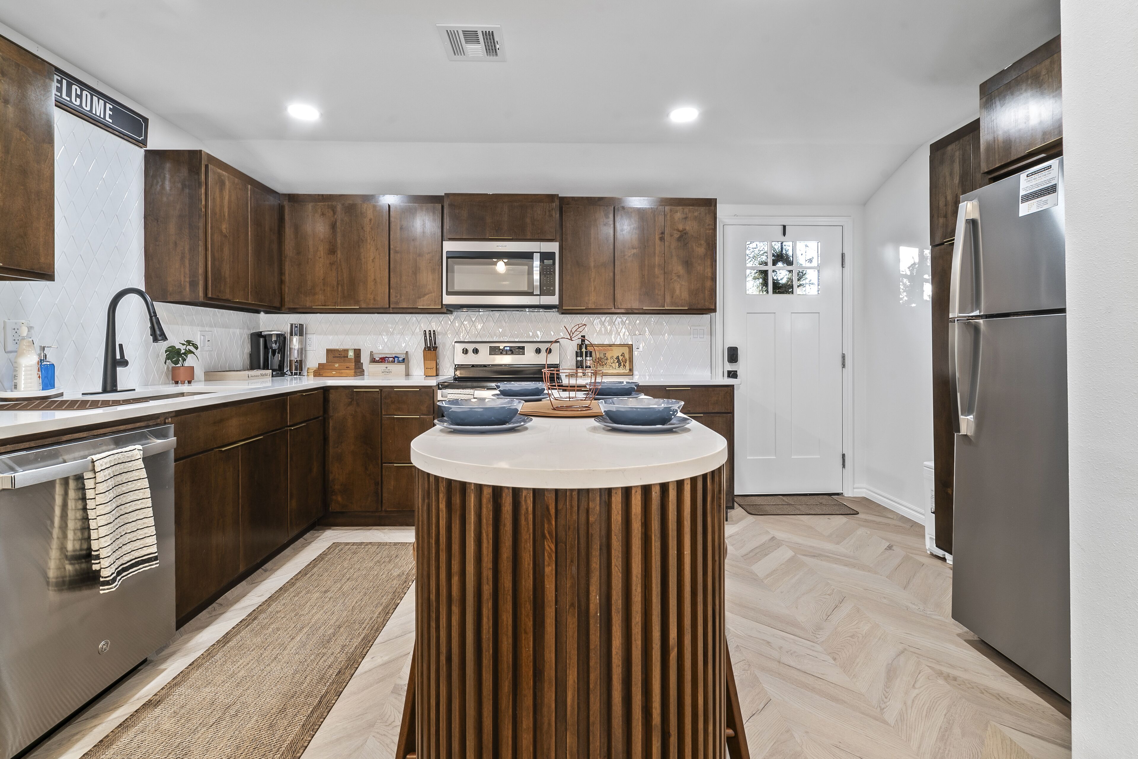 A modern kitchen with wooden cabinets, a central island, stainless steel appliances, and a white tiled backsplash.