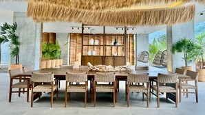 Een eetruimte met een grote houten tafel en rieten stoelen, omringd door potplanten en een modern wandrek met decoratieve objecten.