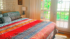 A bedroom with a large bed, a ceiling fan, a window with curtains, and a view of greenery outside.