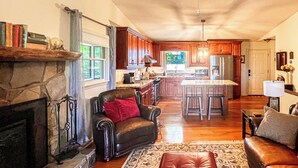 A spacious living room with a fireplace, leather armchair, ottoman, and a kitchen in the background.