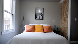 A neatly made bed with white and orange bedding, a framed picture of a bridge on the wall, and a brick accent wall.