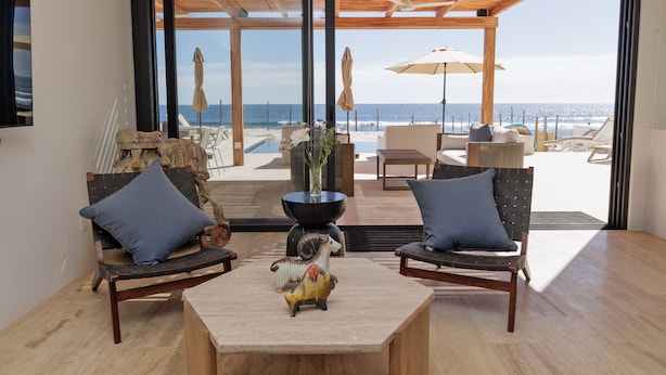 A modern living room with a view of the beach, featuring a sofa, chairs, a coffee table, and a decorative horse figurine. - Exquisite oceanfront luxury casa, Pezula Beach (San Jose del Cabo)