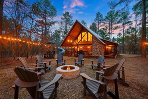 A cabin with a stone exterior and large windows, surrounded by a fire pit and Adirondack chairs, set in a forest.