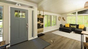Una sala de estar moderna con una puerta gris, un sofá modular negro con almohadas amarillas, una alfombra blanca y ventanas amplias con vistas a zonas verdes.