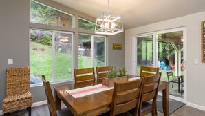 Un comedor con mesa y sillas de madera, ventanas amplias y una lámpara colgante.