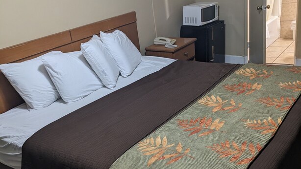 A neatly made bed with white pillows and a brown blanket, a bedside table with a phone and a small appliance, and a view of a bathroom through an open door. - Rainbow Motel (Hendersonville)