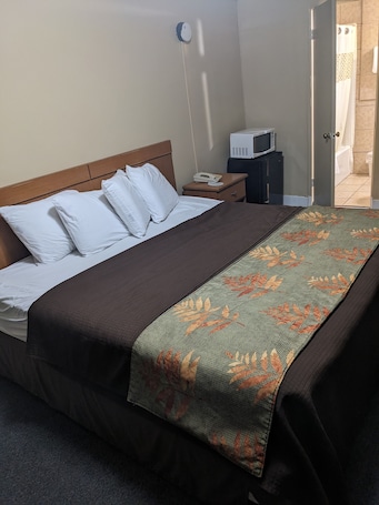A neatly made bed with white pillows and a brown blanket, a bedside table with a phone and a small appliance, and a view of a bathroom through an open door.