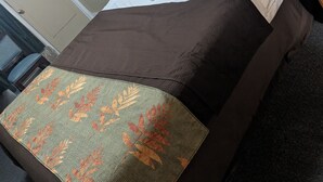 A neatly made bed with white pillows and a brown bedspread, a wooden nightstand with a telephone, and a green leaf-patterned throw blanket on the floor. - Rainbow Motel (Hendersonville)