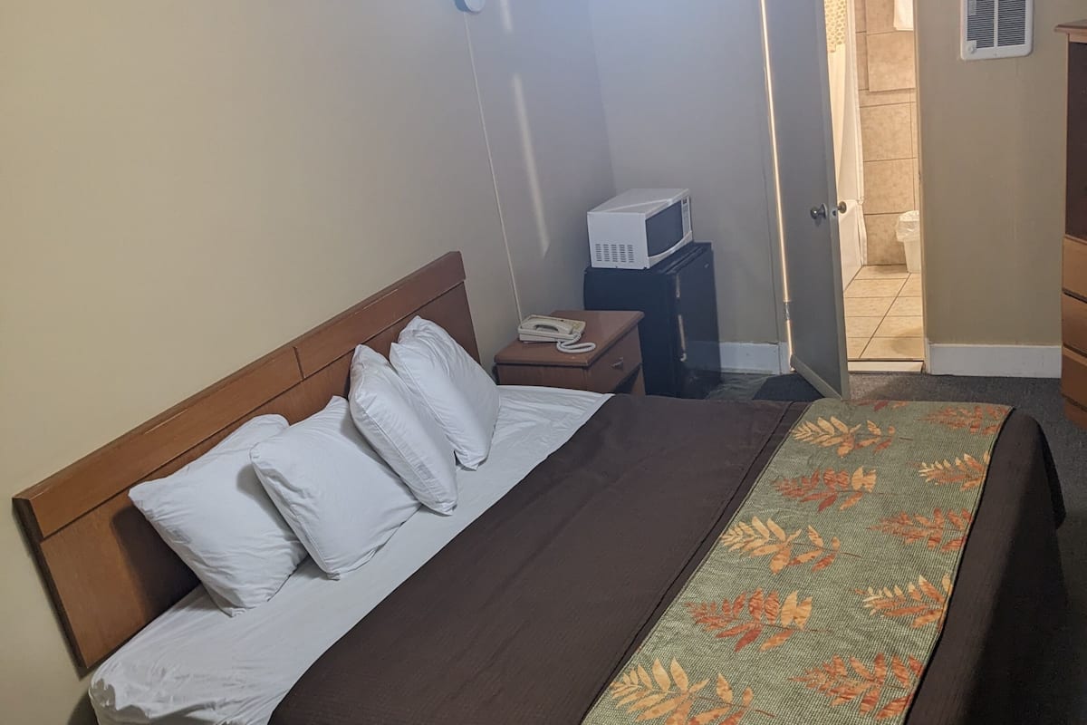 Habitación de hotel con cama, mesita de noche con teléfono y microondas, puerta que da a un baño, y una colcha estampada sobre la cama.