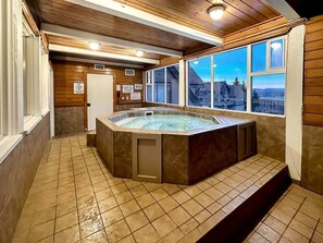 A hot tub in a room with wooden walls and a tiled floor, a window with a view, and a door with a sign. - Slope-Side Serenity Ski-In Ski-Out Gem with Pool (Park City)