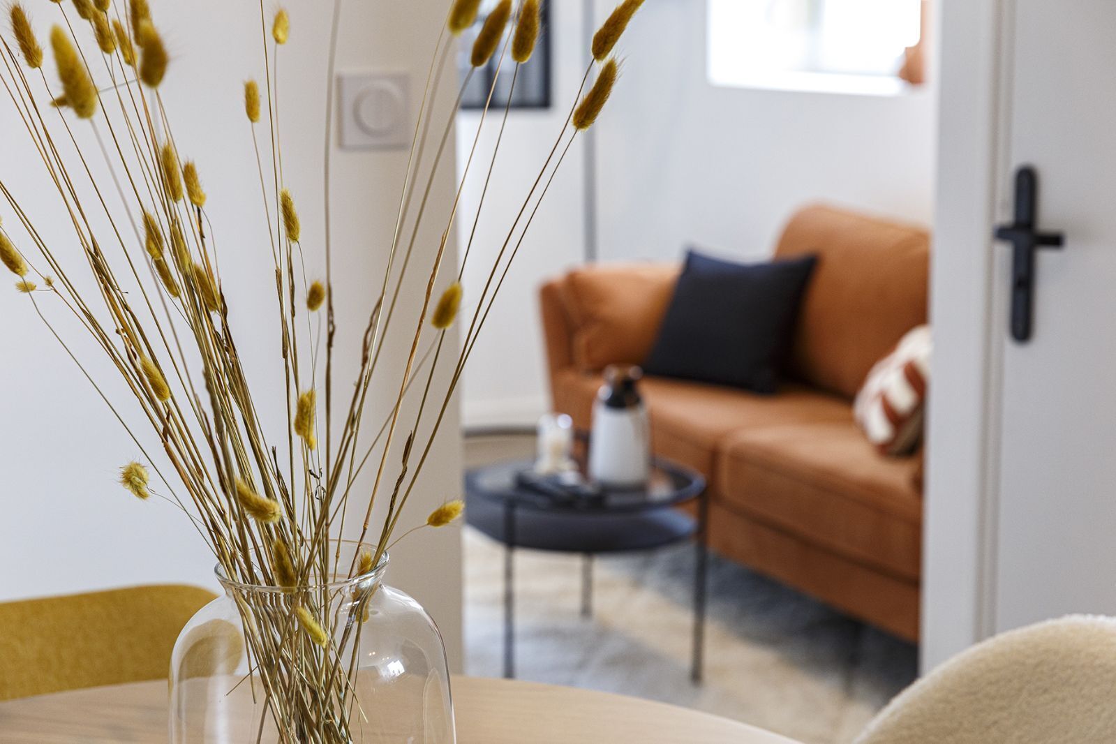A clear view of a vase with dried plants on a table, a brown sofa, and a coffee table with a bottle and a cup in a living room.