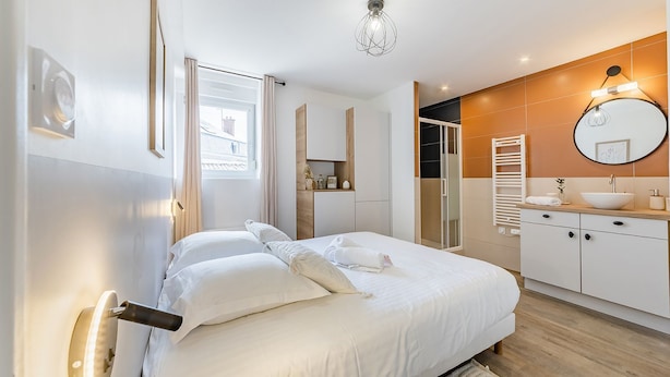 A modern bedroom with a large bed, a bedside table, a built-in wardrobe, a bathroom with a sink and mirror, and a window with curtains. - L'hermès, Douillet et Chaleureux, Venez Vous Détendre (Segré-en-Anjou Bleu)