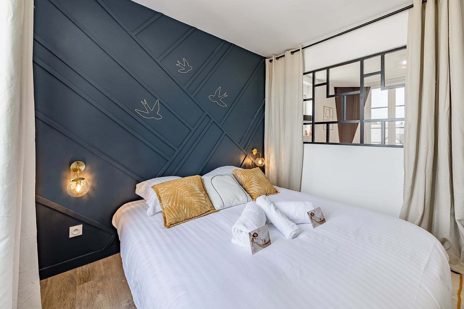 Een hotelkamer met een bed, twee kussens, een nachtkastje, een spiegel en een raam met gordijnen.