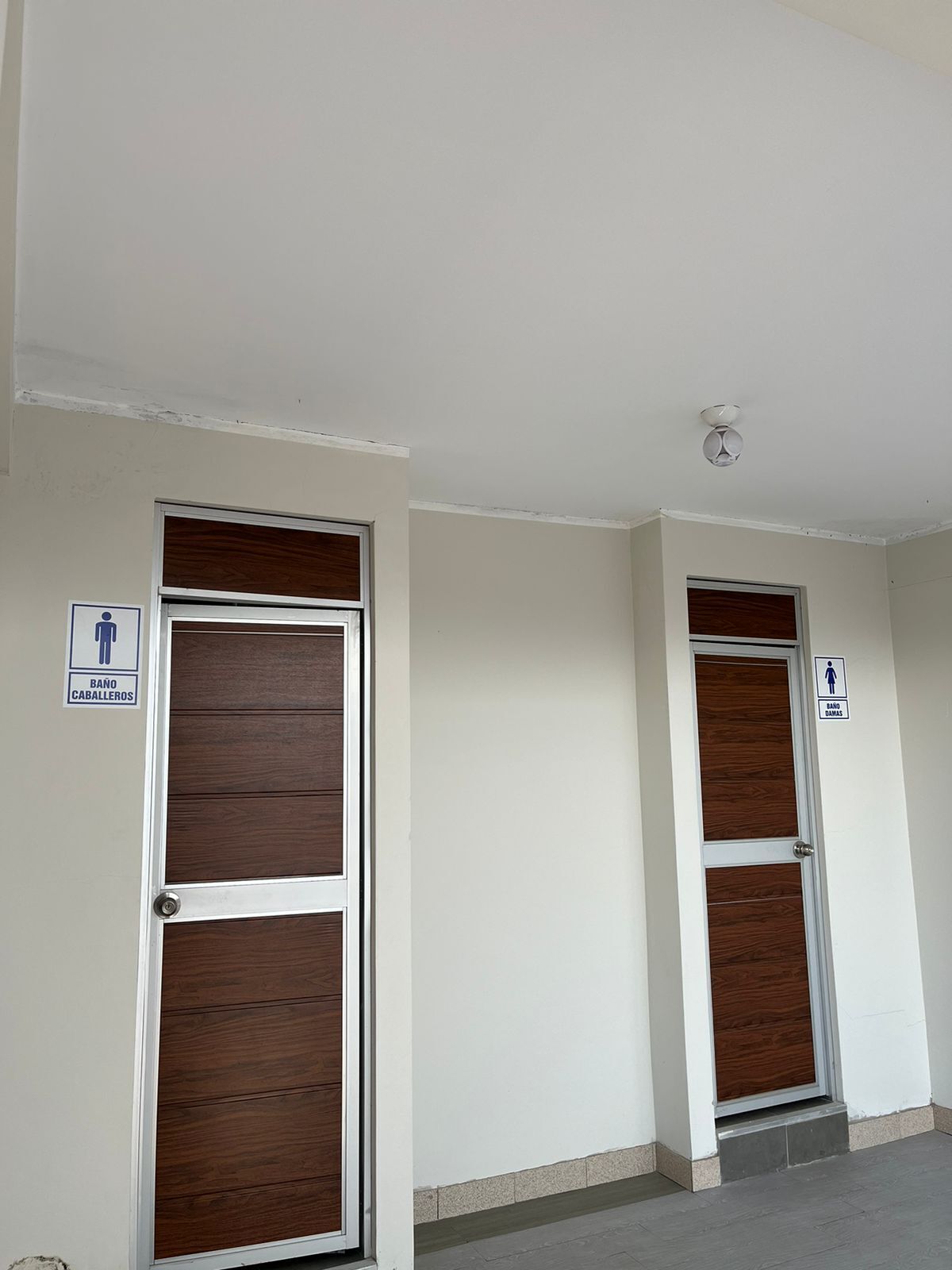 Dos puertas con paneles de madera y marcos metálicos, cada una señalizada con un cartel que indica baños de hombres y de mujeres.